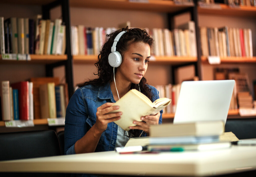 A student refers to a book while working on a laptop with headphones