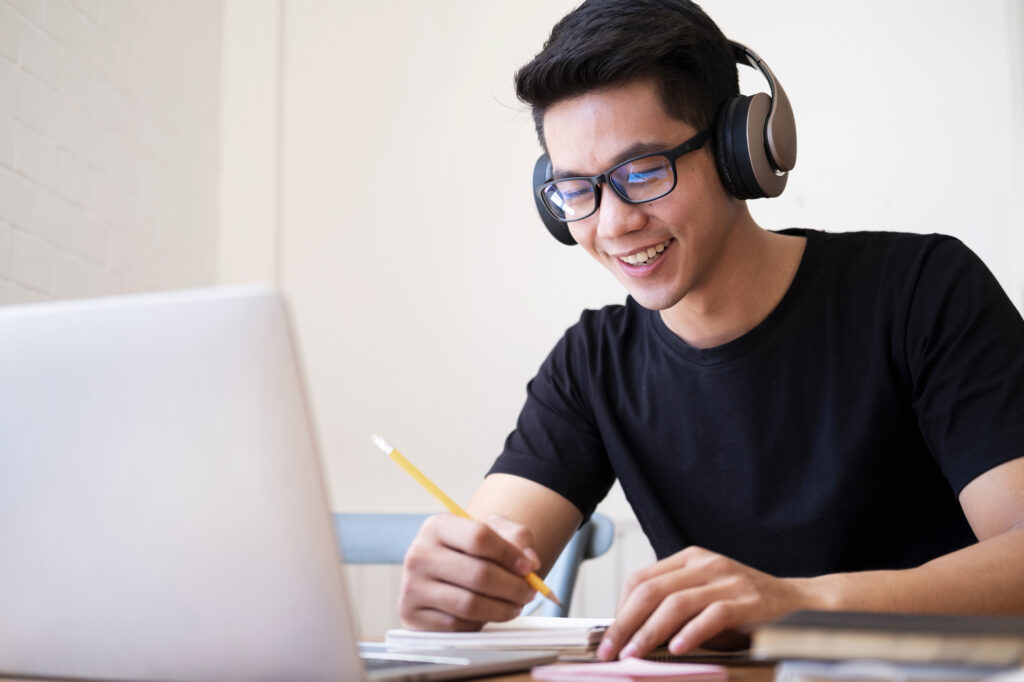 A student smiling while using a laptop and writing notes